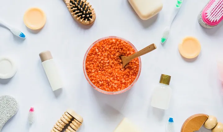 Flat lay of spa essentials including orange bath salts, a brush, toothbrushes, bottles, soaps, and a pumice stone, arranged on a white background.